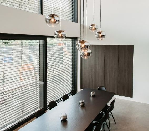 Ein modernes Esszimmer mit einem langen schwarzen Tisch, schwarzen Stühlen, hängenden kugelförmigen Pendelleuchten und großen Fenstern mit Jalousien.