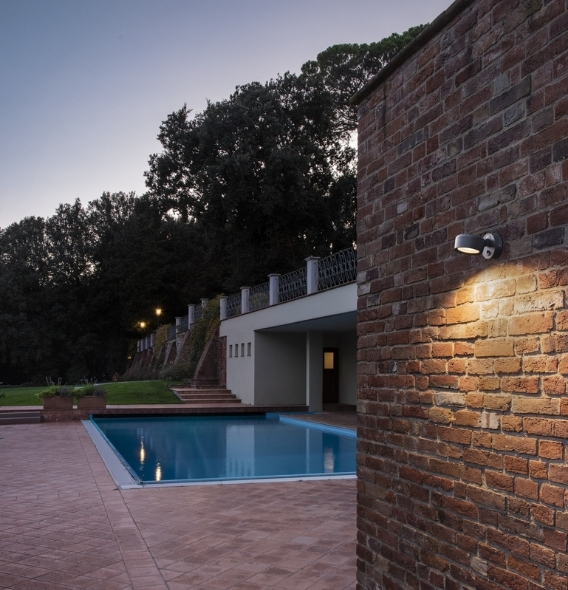 Eine Backsteinmauer mit einer ESKINA Wandleuchte, die in der Abenddämmerung eine Terrasse am Swimmingpool beleuchtet, im Hintergrund sind Bäume zu sehen.