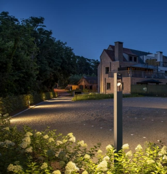 Ein Backsteinhaus mit moderner ESKINA Beleuchtung in der Abenddämmerung, umgeben von Grünflächen und einer gepflasterten Auffahrt, mit Blumen und Bäumen im Vordergrund.