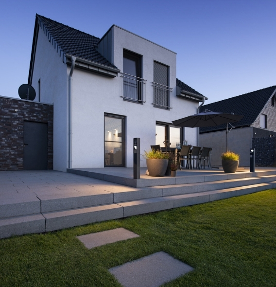 Moderne Terrasse mit Stehleuchten, eleganten Sitzgelegenheiten im Freien, großen Topfpflanzen und einem Sonnenschirm auf stilvoll beleuchteten Stufen. Im Hintergrund ein modernes zweistöckiges Haus.