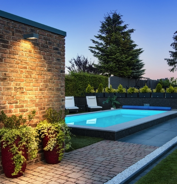 Beleuchteter Garten mit einem kleinen Pool, Liegestühlen und Topfpflanzen. Eine Backsteinmauer und Bäume sorgen für eine ruhige Abendstimmung.