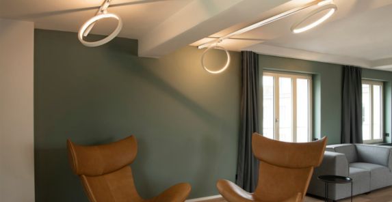 Modernes Wohnzimmer mit braunen Ledersesseln, ONE 40 TRACK DALI Schienenleuchten, grünen Wänden, großen Fenstern mit dunklen Vorhängen und einem grauen Sofa.