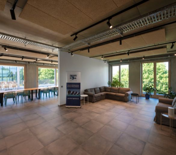 Geräumiger Raum mit großem Sofa, Tischen, Stühlen und Topfpflanzen. Große Fenster bieten einen Blick auf die grüne Umgebung. NUMINOS® Deckenspots beleuchten den Raum.