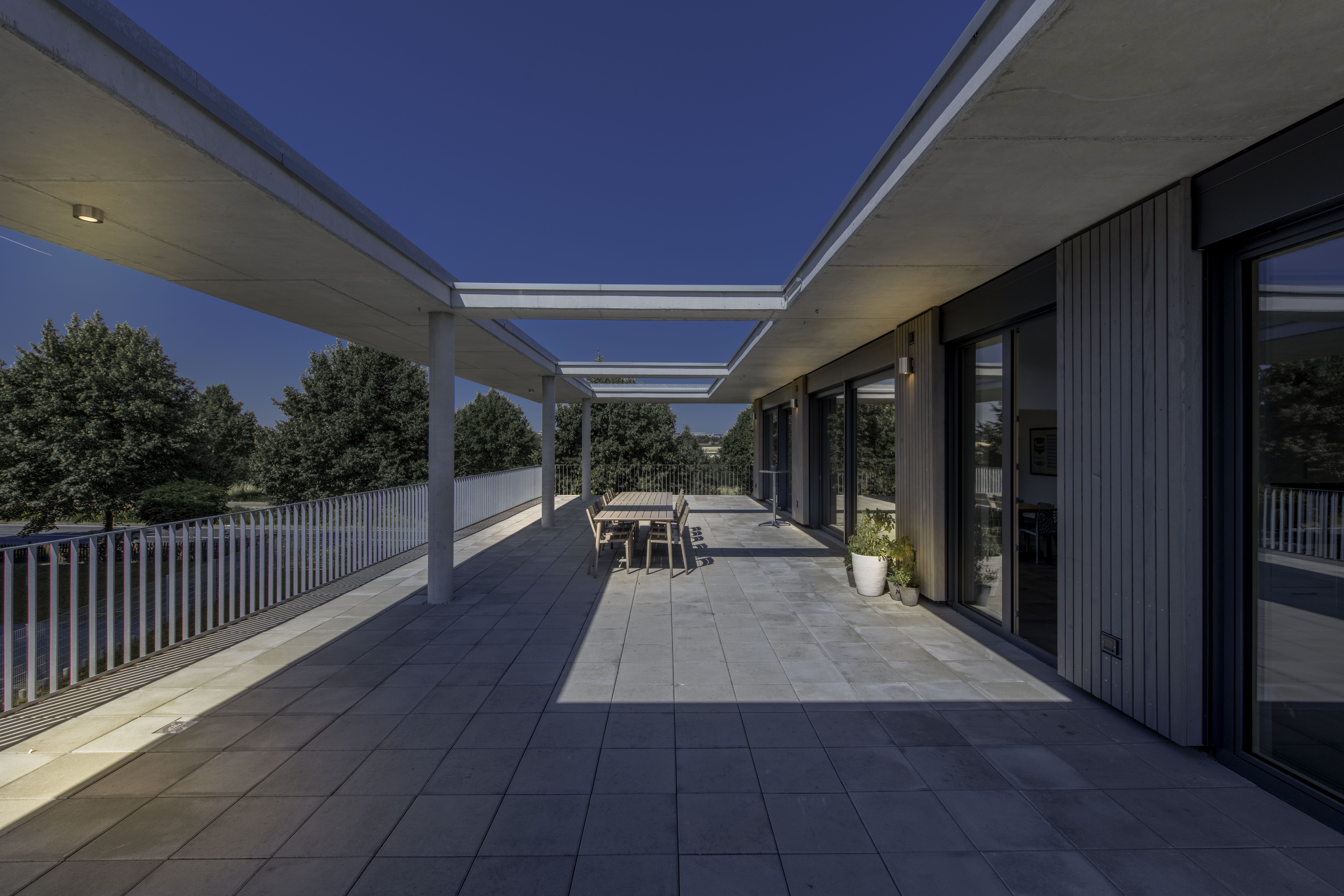 Geräumige Dachterrasse mit Fliesenboden, einem langen Esstisch und einem Geländer unter strahlend blauem Himmel - beleuchtet von ROX UP/DOWN Wandleuchten. Im Hintergrund sind Bäume zu sehen.