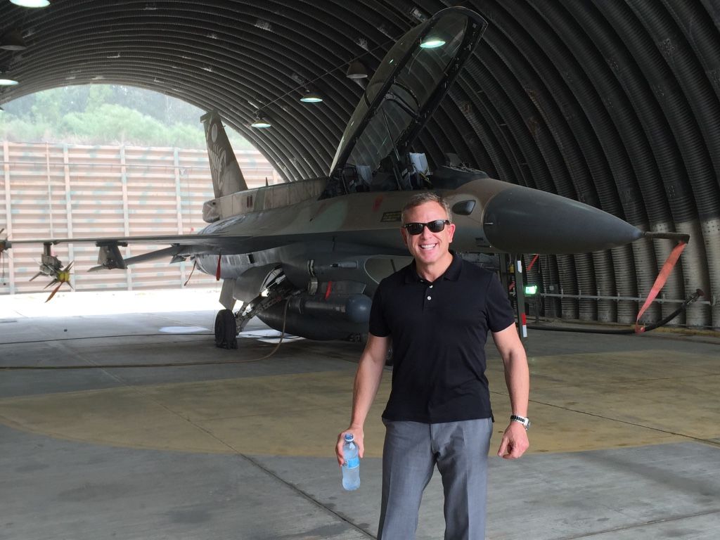 Brett Kingstone in sunglasses holding a water bottle stands in front of a fighter jet inside a hangar.