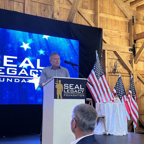 Brett Kingstone speaking at a podium at the Max King Ranch in Colorado with "SEAL Legacy Foundation" signage, in a wooden venue with American flags and a large screen behind him.