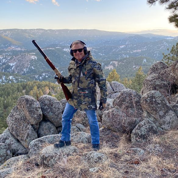 Brett Kingstone at the Max King Ranch standing on a rocky environment wearing hunting gear and holding a hunting rifle.