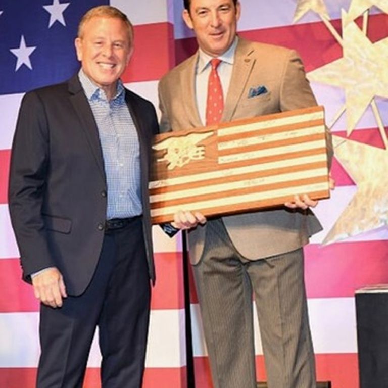 Two men in suits stand on stage; one holds a wooden American flag plaque. The backdrop features stars and stripes.