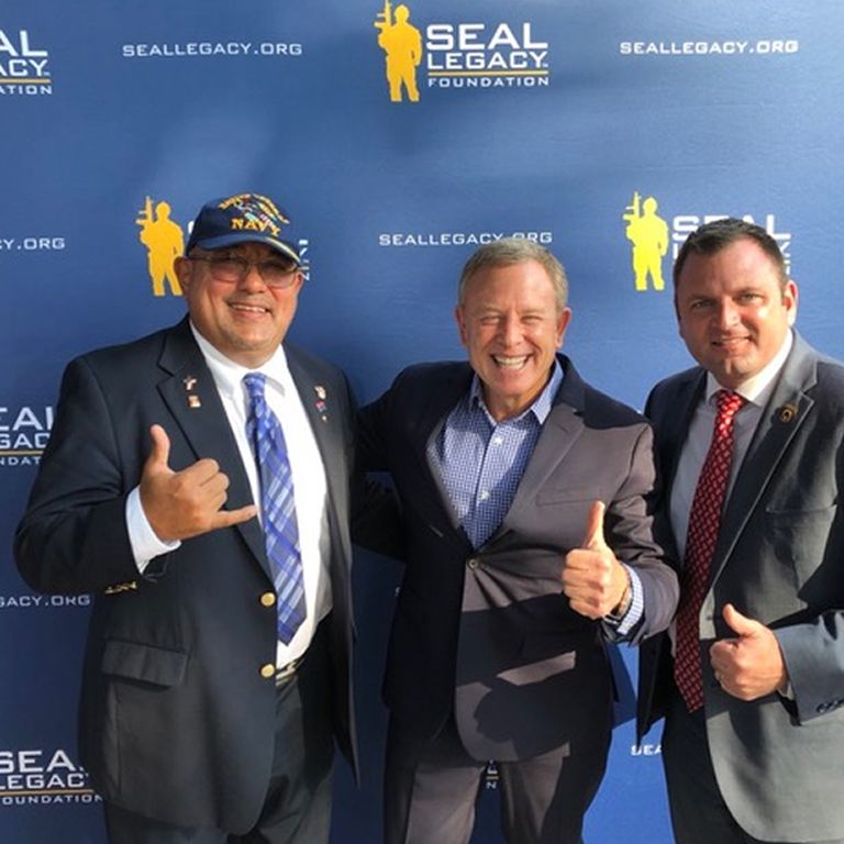 Three men smiling and posing in front of a blue SEAL Legacy Foundation backdrop, two giving thumbs-up gestures.