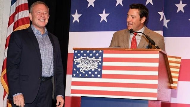 Two men at a podium with a U.S. flag design; one speaks at a microphone while the other smiles. An American flag is visible in the background.