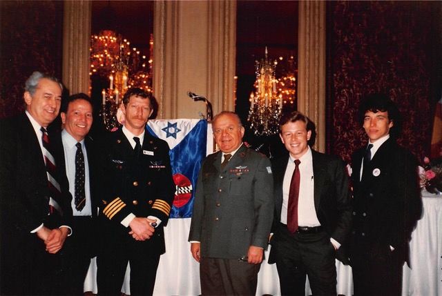 A group of six men, including military personnel, stand smiling in a formal setting with a flag and microphone in the background.
