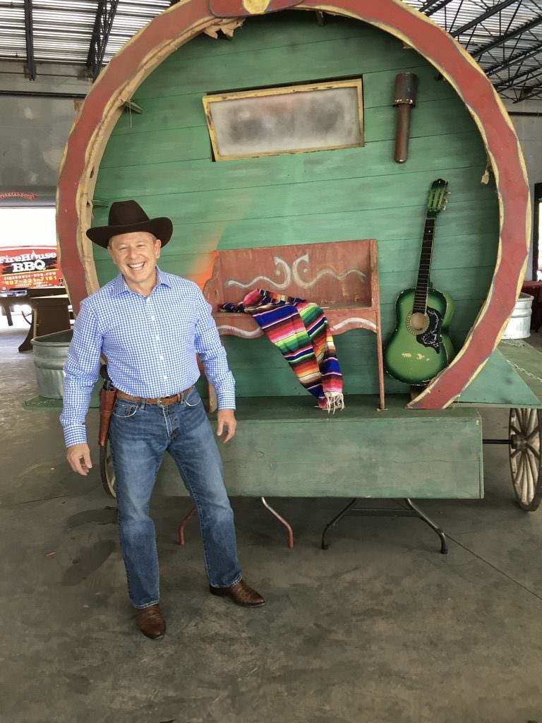 Brett Kingstone in cowboy hat and blue checkered shirt stands smiling beside a colorful wagon with a guitar and blanket inside.