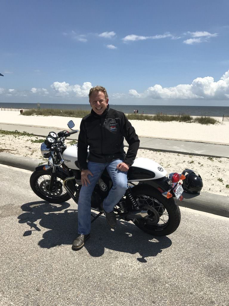 Brett Kingstone sitting on a motorcycle parked by a beach, wearing a black jacket and jeans. The sky is clear with a few clouds.
