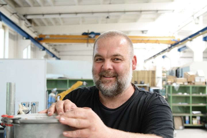 Un dipendente della Woehler è in piedi davanti a un macchinario nel capannone di produzione, con un componente tra le mani. Sullo sfondo si vedono le attrezzature industriali e i sistemi di scaffalature di un moderno ambiente di produzione.