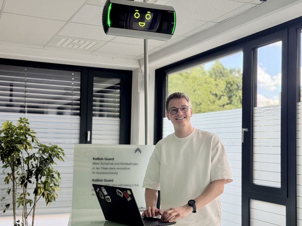 Ein Mitarbeiter steht lächelnd mit einem Laptop unter dem an der Decke montierten KeBob Guard. Das Gerät zeigt ein stilisiertes Gesicht mit grün leuchtenden Elementen.