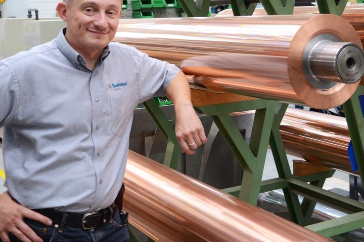 Hansjörg Michel, Head of Technology bei Daetwyler Graphics steht neben großen, glänzenden Kupferrollen auf einem grünen Industrieregal.