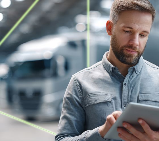 Mann arbeitet mit einem Tablet in moderner Industrieumgebung; im Hintergrund sind Lastwagen und digitale Linien sichtbar.