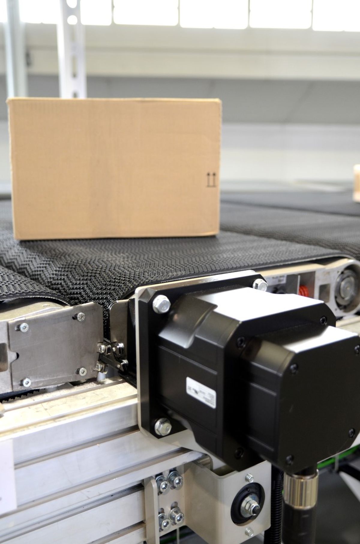 Primo piano di una scatola di cartone su un nastro trasportatore automatizzato con unità di azionamento in primo piano – simbolo di movimentazione precisa dei materiali e automazione industriale nella logistica.