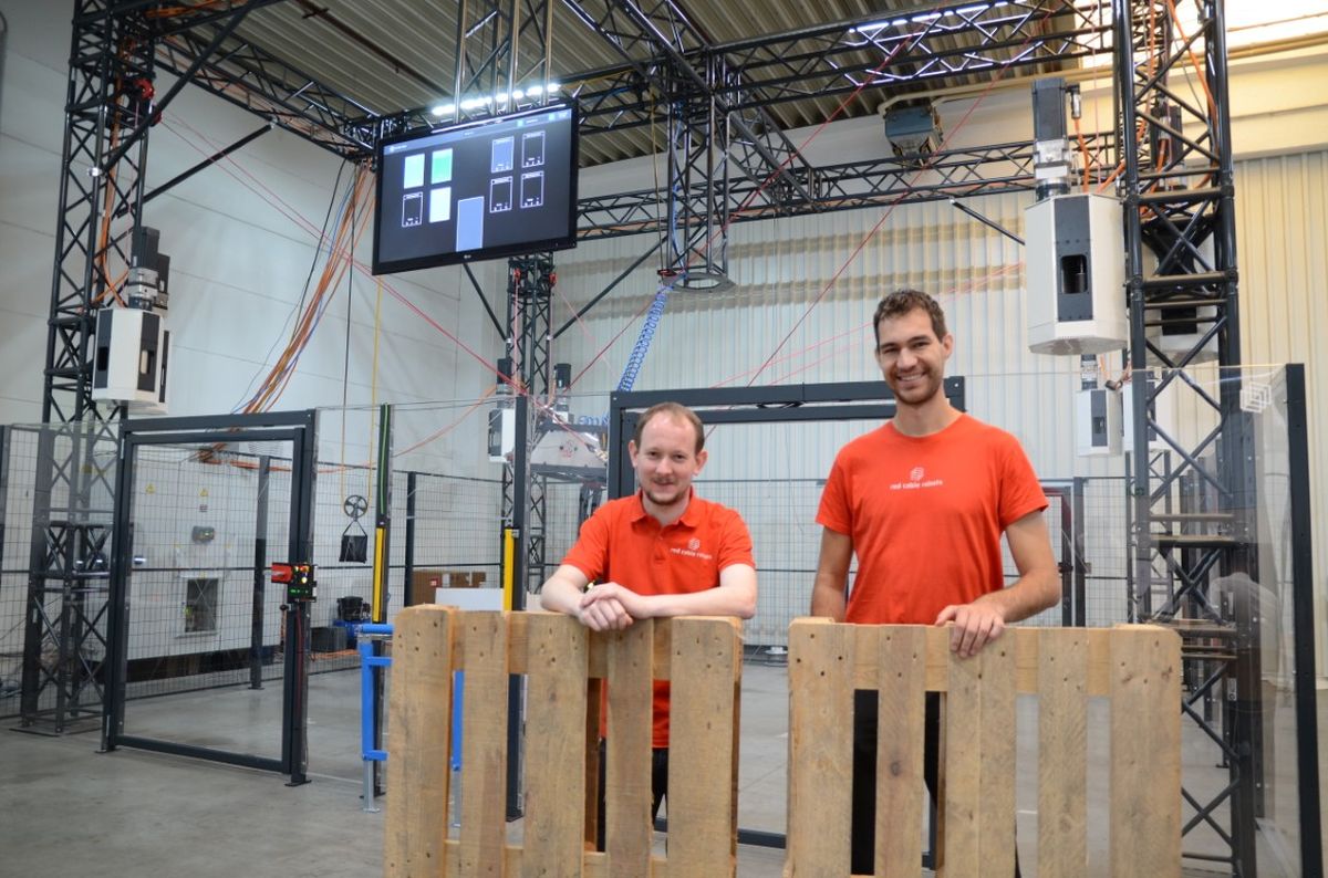 Patrik Lemmen (CTO) and Robin Heidel (COO) of Red Cable Robots stand in an industrial hall in front of a cable robot system. The image conveys expertise in industrial automation and the close link between technology and company leadership.