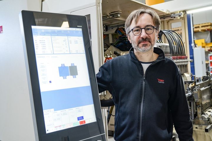 Stefan Cämmerer, Head of Software Development and Electrical Design, stands next to an HMI panel and looks at the camera, while a machine is visible in the background. The display shows graphical representations and process data.