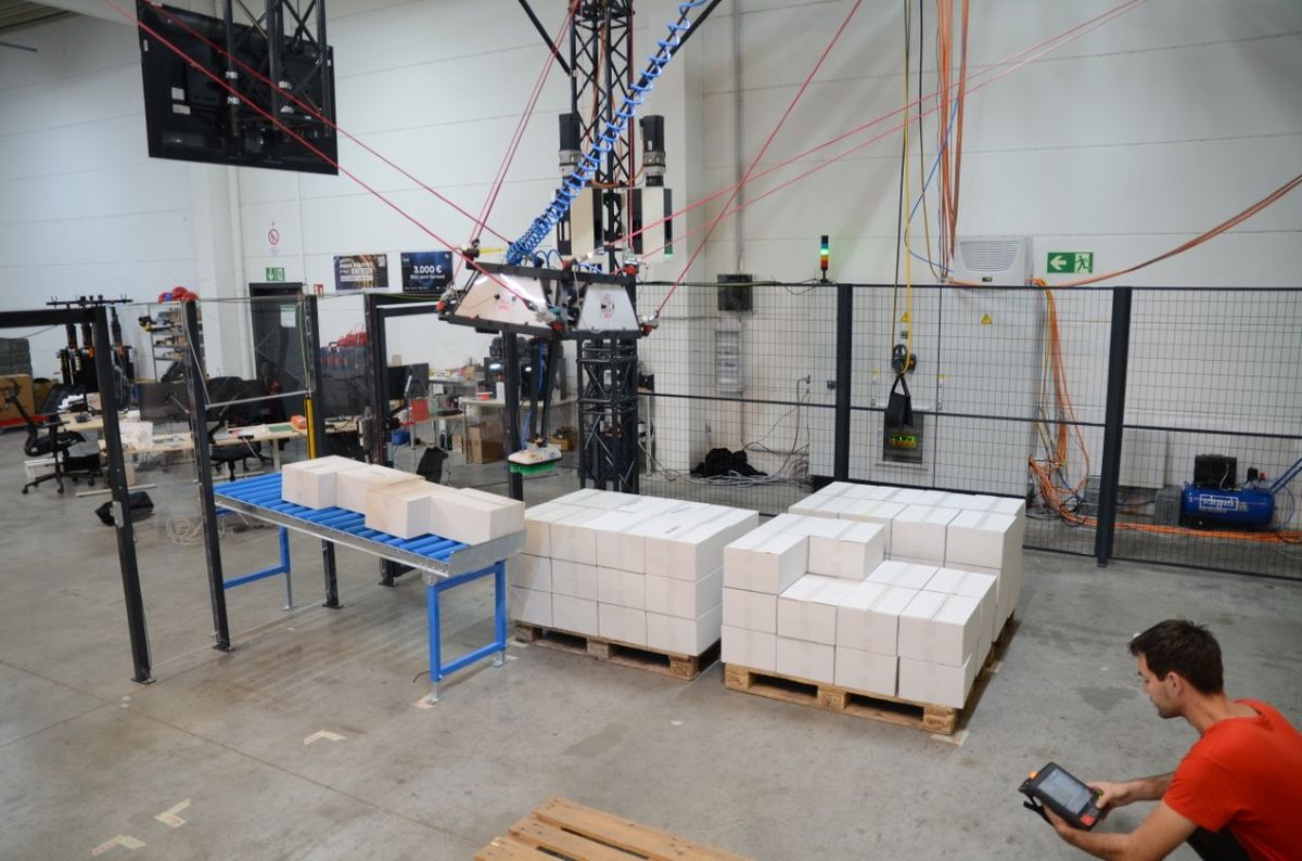 A cable robot operates above pallets with boxes in an industrial hall while an operator uses a wireless control device at a distance. The image highlights flexible and safe robot teaching and monitoring enabled by wireless HMI.