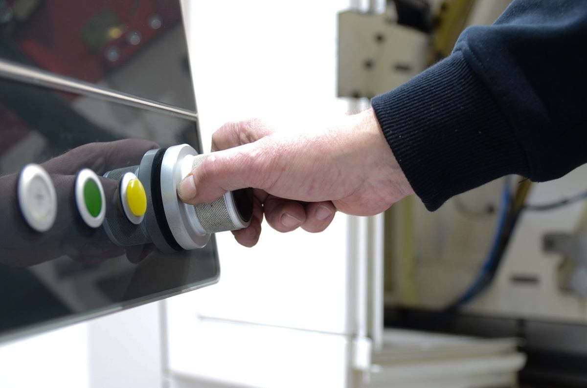 A hand operates a rotary knob next to colored buttons on a machine panel. This illustrates a physical input method for machine control.