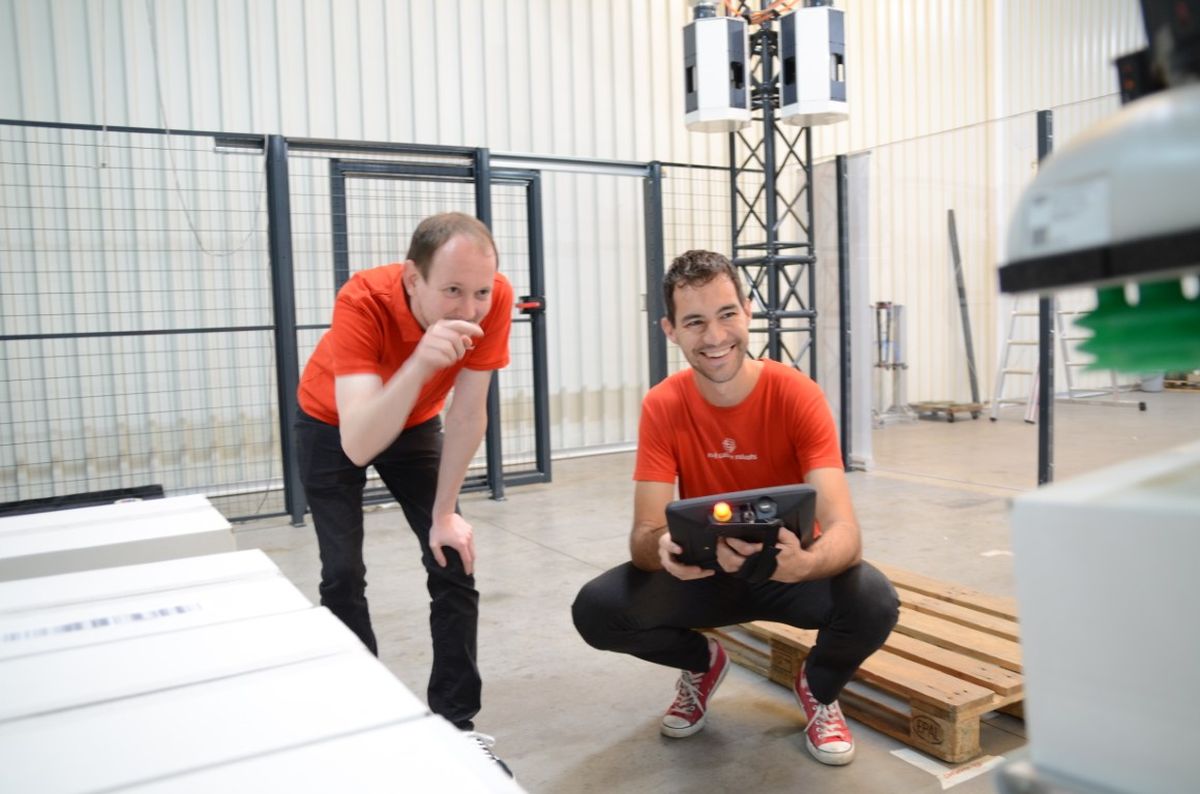 Patrik Lemmen (CTO) and Robin Heidel (COO) of Red Cable Robots work in an industrial hall on an automated cable robot system. One operates a control device while both observe the material handling process.