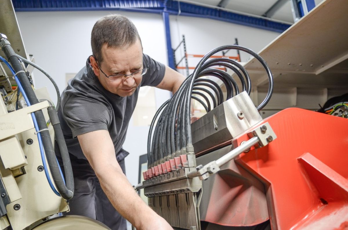 A man is operating industrial machinery and adjusting the controls in a large workshop. He is wearing glasses and a gray shirt.