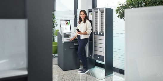 A woman in a blue top is smiling as she stands next to a KEBA self-service terminal in a modern office with a green chair and plants.