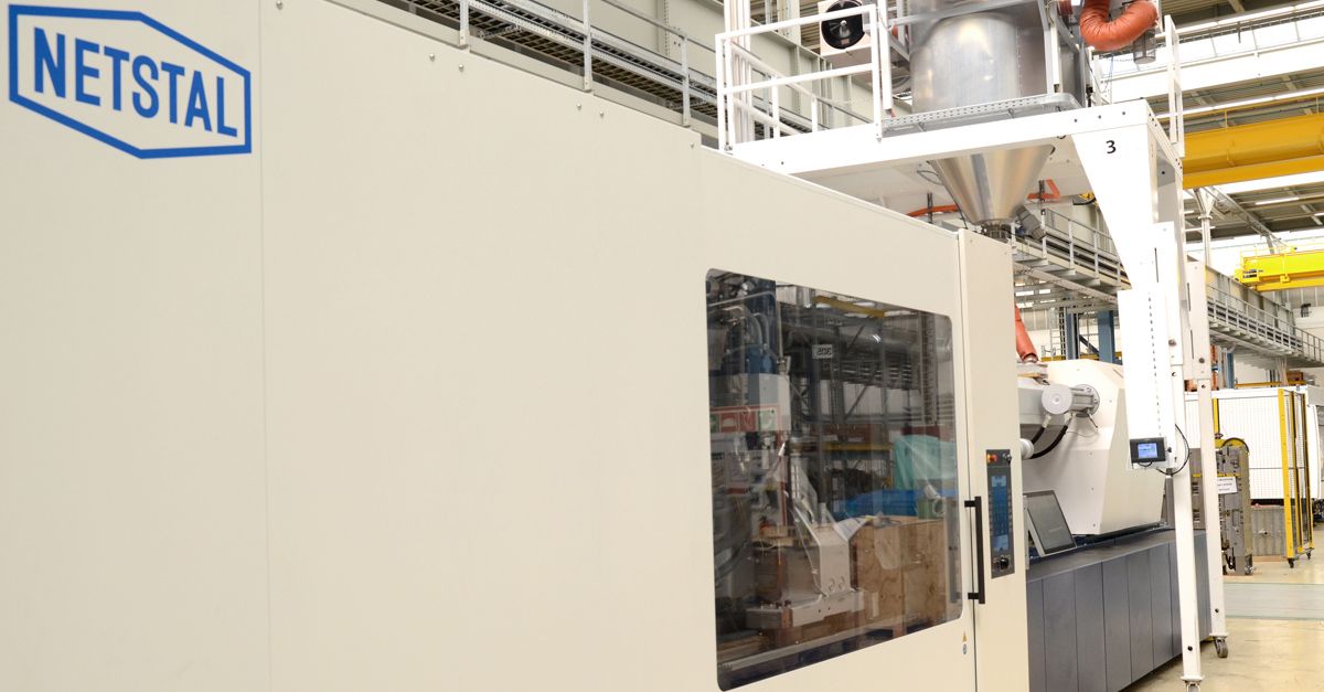 Macchina per stampaggio a iniezione NETSTAL in un moderno stabilimento di produzione. Sopra la macchina è visibile un sistema di alimentazione con tramoggia e tubazioni, simbolo della lavorazione automatizzata della plastica e del controllo preciso.