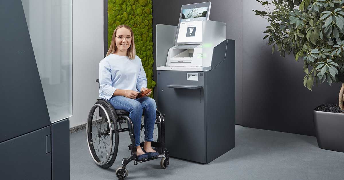 A woman in a wheelchair is sitting in front of a modern self-service terminal in an accessible bank branch.