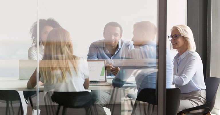 A diverse group of professionals sitting around a table in a glass meeting room, engaged in a discussion. A laptop with a presentation open is visible on the table, suggesting a collaborative work or training session.