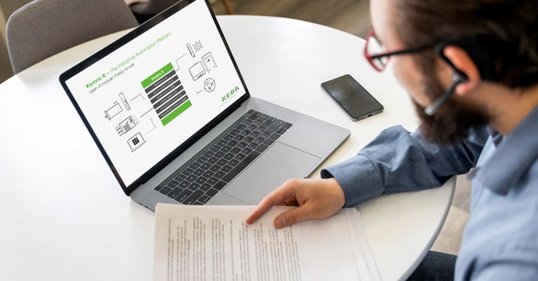 Ein Mann mit Kopfhörern sieht sich an einem Tisch ein Dokument an, neben ihm stehen ein Laptop, auf dem ein Web-Seminar von KEBA Industrial Automation läuft.