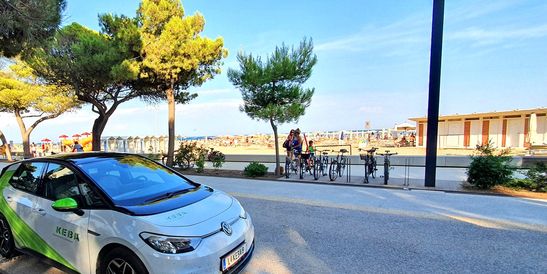 A white and green electric car from KEBA is parked on a road near the beach, surrounded by palm trees and bicycles. In the background, the sea and a bathing area are visible – holiday vibes with sustainable mobility.