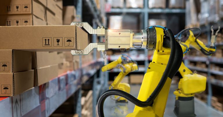 Gelber Roboterarm, der einen Karton in einem Lagerhaus handhabt, im Hintergrund Regale mit gestapelten Kartons.