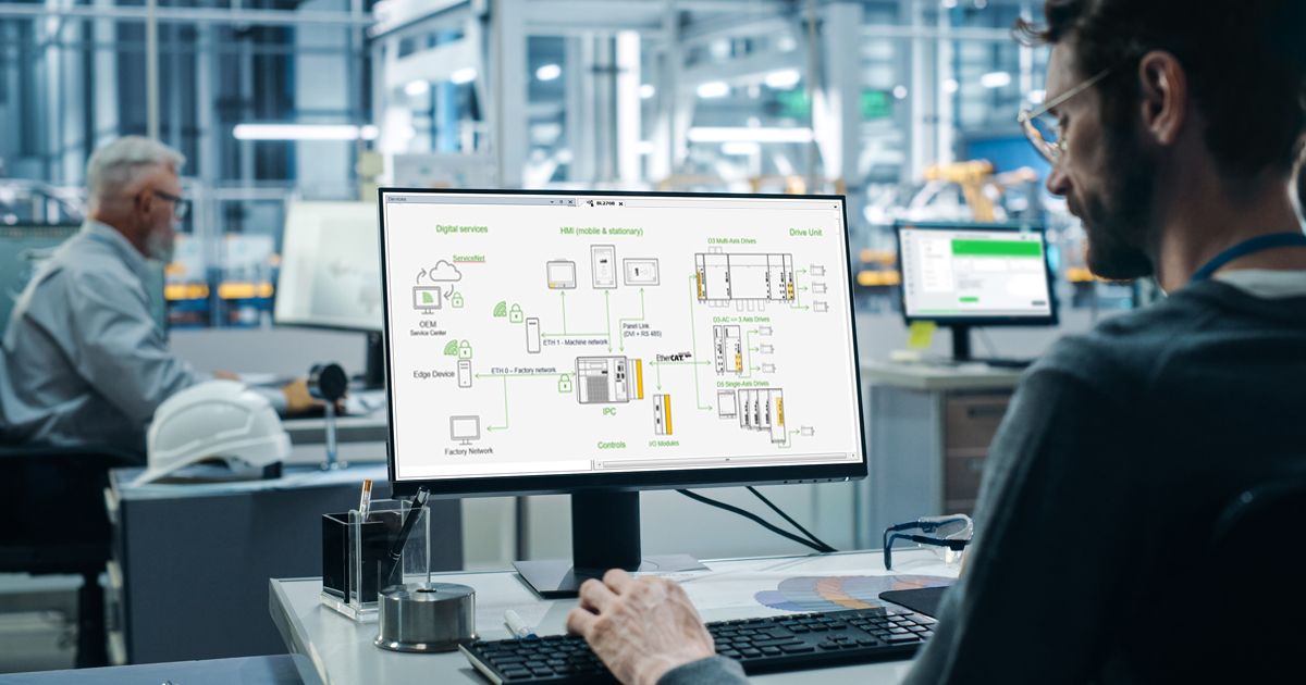 An engineer sits in front of a computer screen in a modern industrial environment and analyzes a graphical diagram of an automation system (Kemro X). The monitor displays various devices, controllers, and network elements connected to each other. In the background, another colleague works in an industrial production environment.