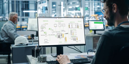 An engineer sits in front of a computer screen in a modern industrial environment and analyzes a graphical diagram of an automation system (Kemro X). The monitor displays various devices, controllers, and network elements connected to each other. In the background, another colleague works in an industrial production environment.