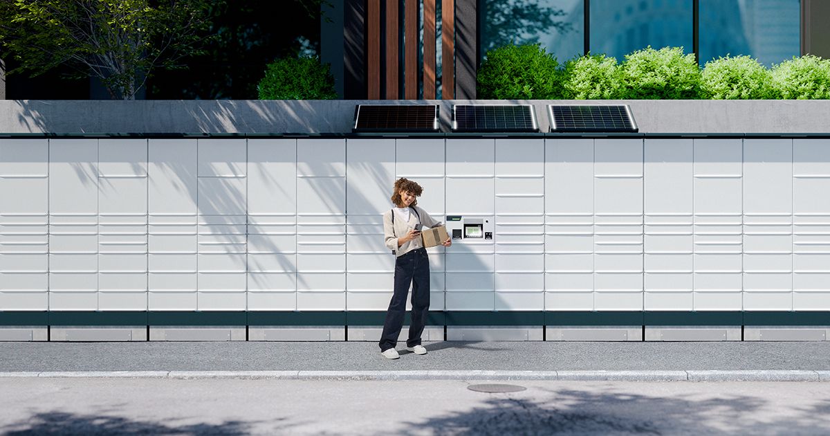 Paketstation im Außenbereich einer modernen Wohngegend mit Pflanzen und Gebäuden im Hintergrund.