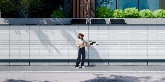 Paketstation im Außenbereich einer modernen Wohngegend mit Pflanzen und Gebäuden im Hintergrund.