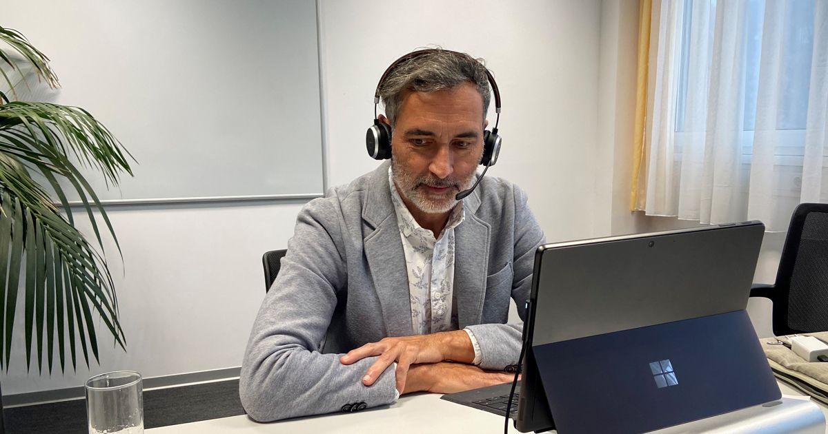 Man with headset sitting in front of his laptop at a desk, in an office with a plant, whiteboard, and window curtain.
