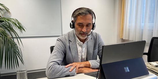 Man with headset sitting in front of his laptop at a desk, in an office with a plant, whiteboard, and window curtain.