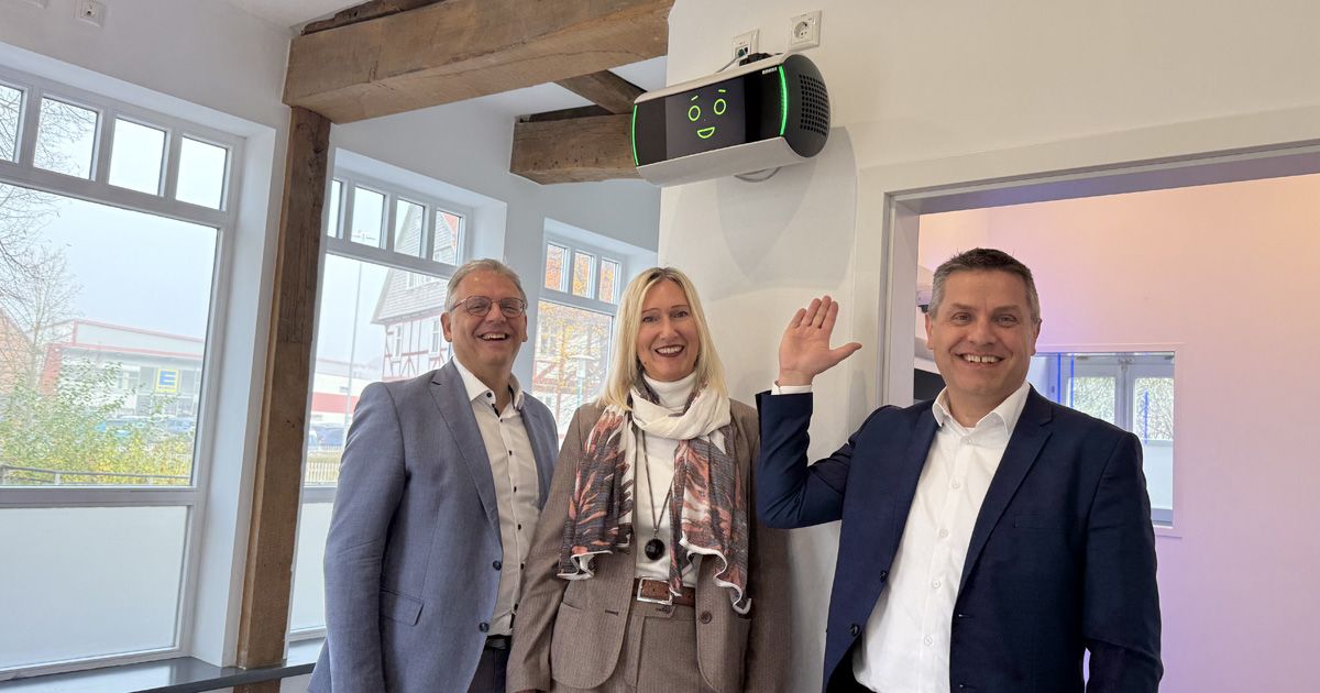 Drei lächelnde Fachleute in einem Bankfoyer zeigen auf ein an der Wand montiertes KI-Assistenzsystem mit grünen Anzeigeleuchten.