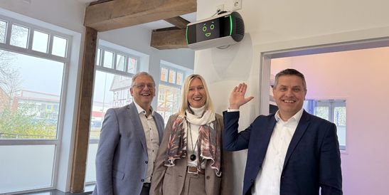 Drei lächelnde Fachleute in einem Bankfoyer zeigen auf ein an der Wand montiertes KI-Assistenzsystem mit grünen Anzeigeleuchten.