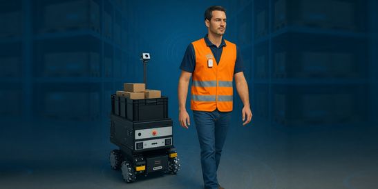 Ein Lagerarbeiter mit orangefarbener Sicherheitsweste geht neben einem autonomen mobilen Roboter, der gestapelte Kartons in einer modernen Lagerumgebung transportiert. environment.
