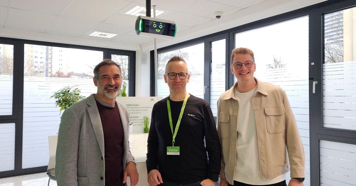 Three people standing indoors in front of an AI based assistance system, smiling. One wears a gray blazer, another a black shirt with a green lanyard, and the third a beige jacket.