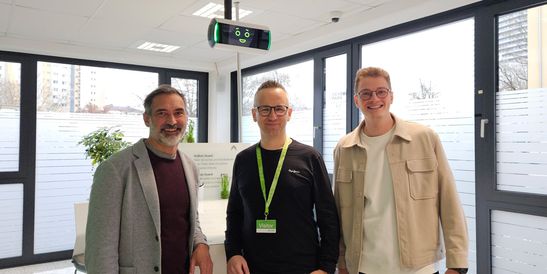Three people standing indoors in front of an AI based assistance system, smiling. One wears a gray blazer, another a black shirt with a green lanyard, and the third a beige jacket.
