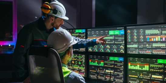 Two industrial workers wearing safety helmets monitor multiple screens with complex data visualizations in a control center.