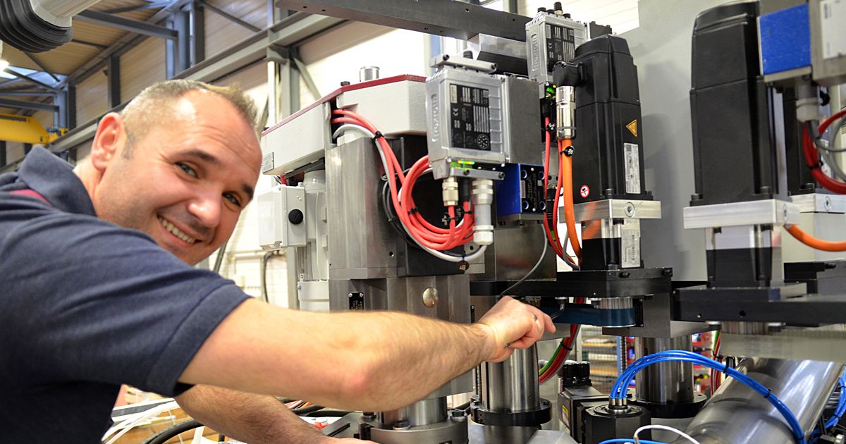 Service engineer setting up a complex industrial machine with drives, sensors, and wiring, smiling at the camera. The image represents expertise, precision, and enthusiasm in industrial automation work.