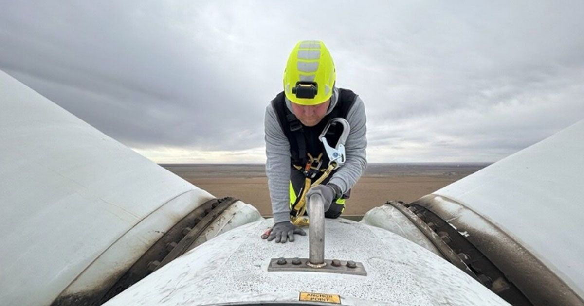 Ein Techniker mit Sicherheitsausrüstung arbeitet auf einer Windkraftanlage zwischen zwei Rotorblättern unter bewölktem Himmel. Das Bild zeigt Wartungs- und Servicearbeiten im Bereich erneuerbare Energien und industrielle Infrastruktur