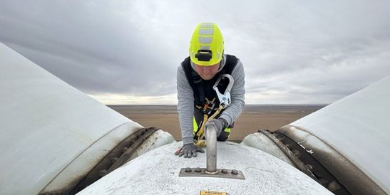 Ein Techniker mit Sicherheitsausrüstung arbeitet auf einer Windkraftanlage zwischen zwei Rotorblättern unter bewölktem Himmel. Das Bild zeigt Wartungs- und Servicearbeiten im Bereich erneuerbare Energien und industrielle Infrastruktur